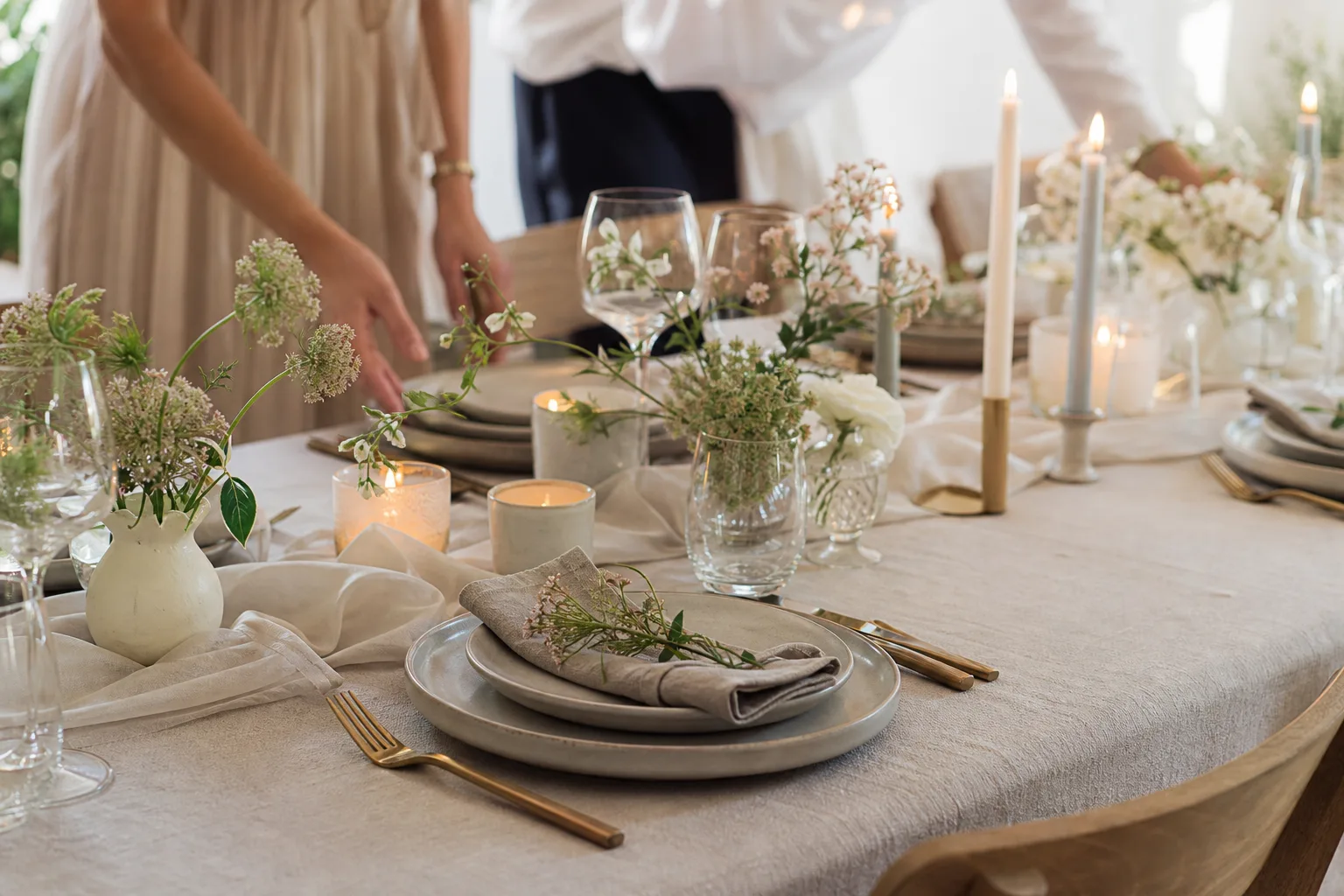 Ett dukat bröllopsbord med linne, porslin, ljus och enkla blommor.